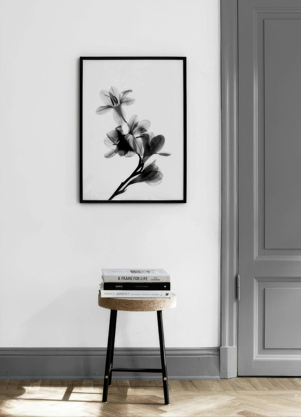 Affiche noir et blanc de fleurs translucides, suspendue dans une pièce minimaliste avec un tabouret et des livres.