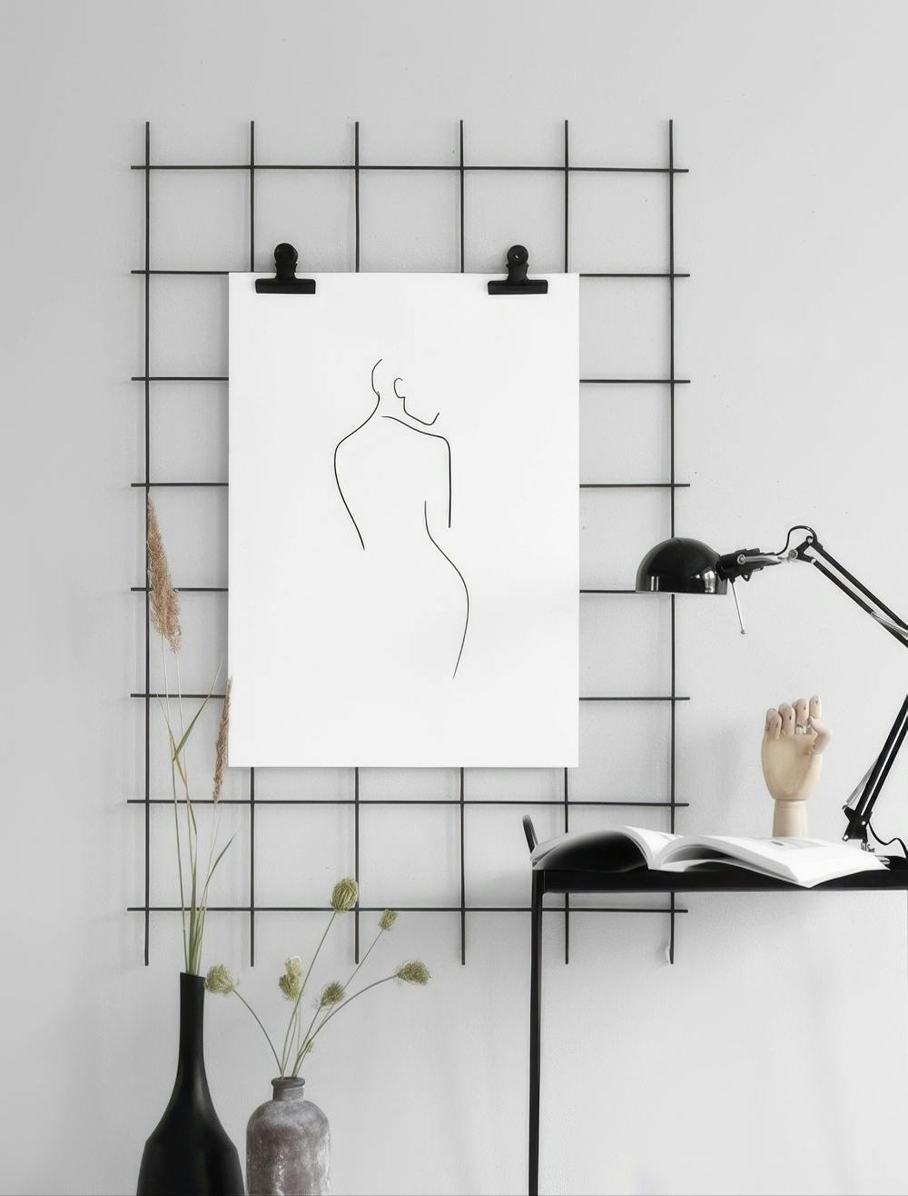 Affiche dune silhouette féminine en trait continu noir, suspendue sur une grille murale dans un intérieur moderne.