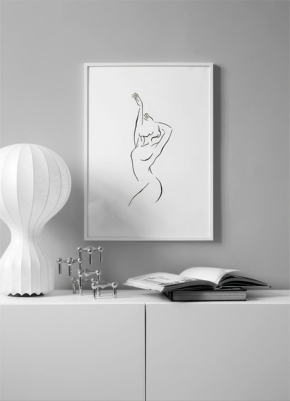 Affiche dune silhouette féminine minimaliste en noir et blanc, suspendue au-dessus dun meuble blanc avec lampe et livres.