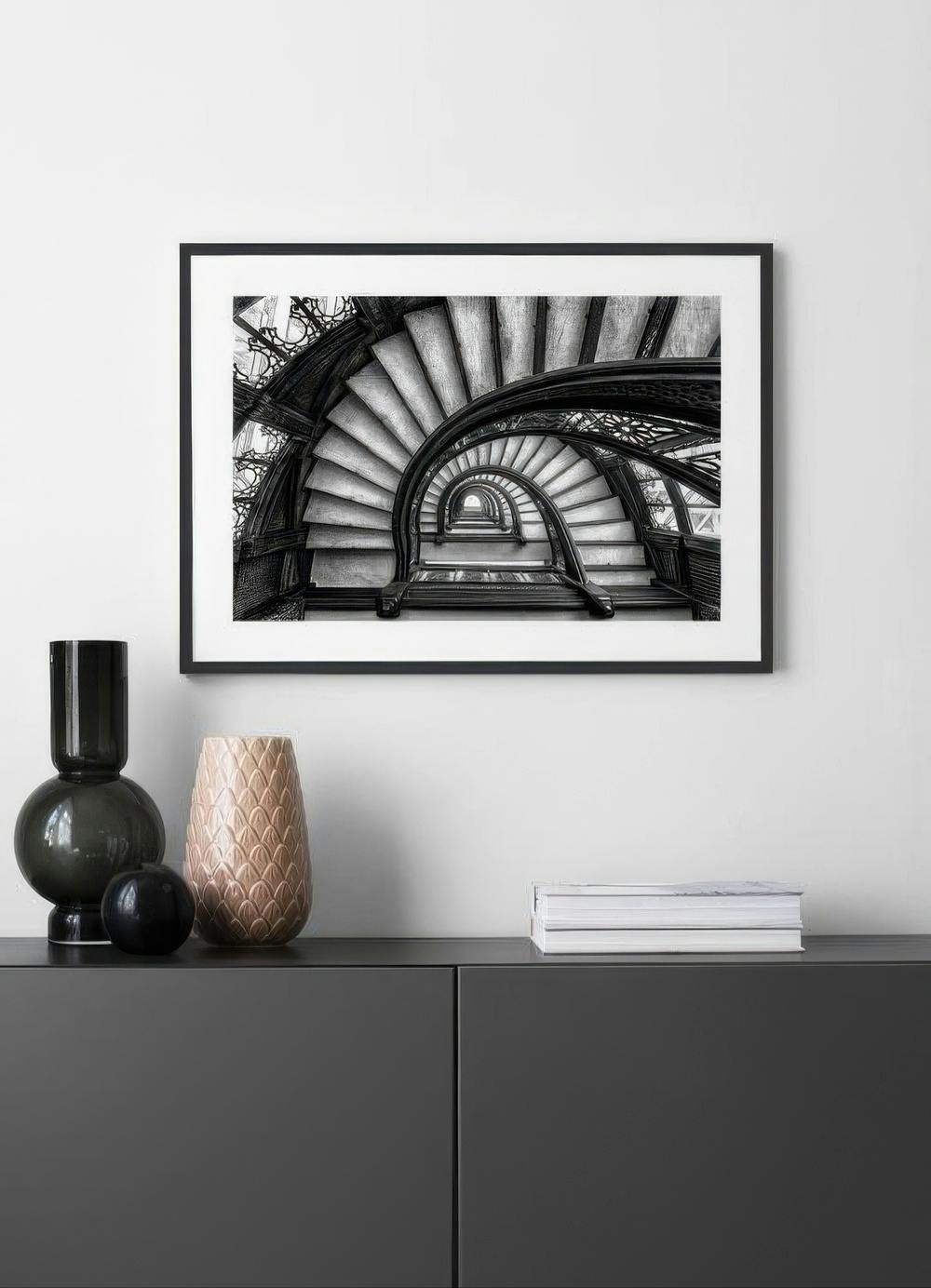 Affiche en noir et blanc dun escalier en colimaçon, suspendue au-dessus dun meuble bas avec deux vases et des livres.