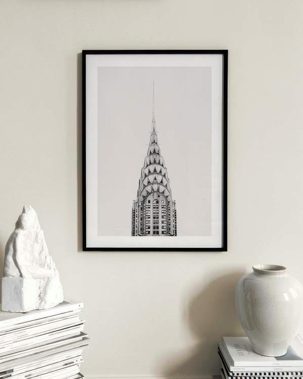 Affiche du Chrysler Building de New York en noir et blanc, accrochée au mur au-dessus dune pile de magazines et dun vase.