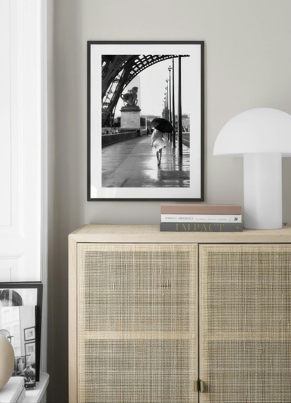 Affiche noir et blanc d’une femme sous parapluie sur un pont de Paris, suspendue au-dessus d’un buffet en rotin.