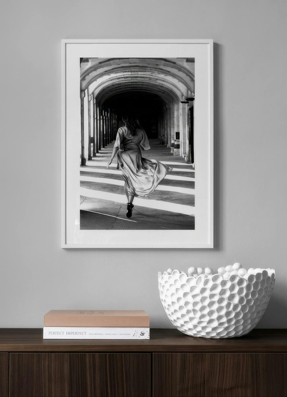 Affiche en noir et blanc dune femme en robe fluide marchant dans une arche, suspendue au-dessus dun meuble en bois.