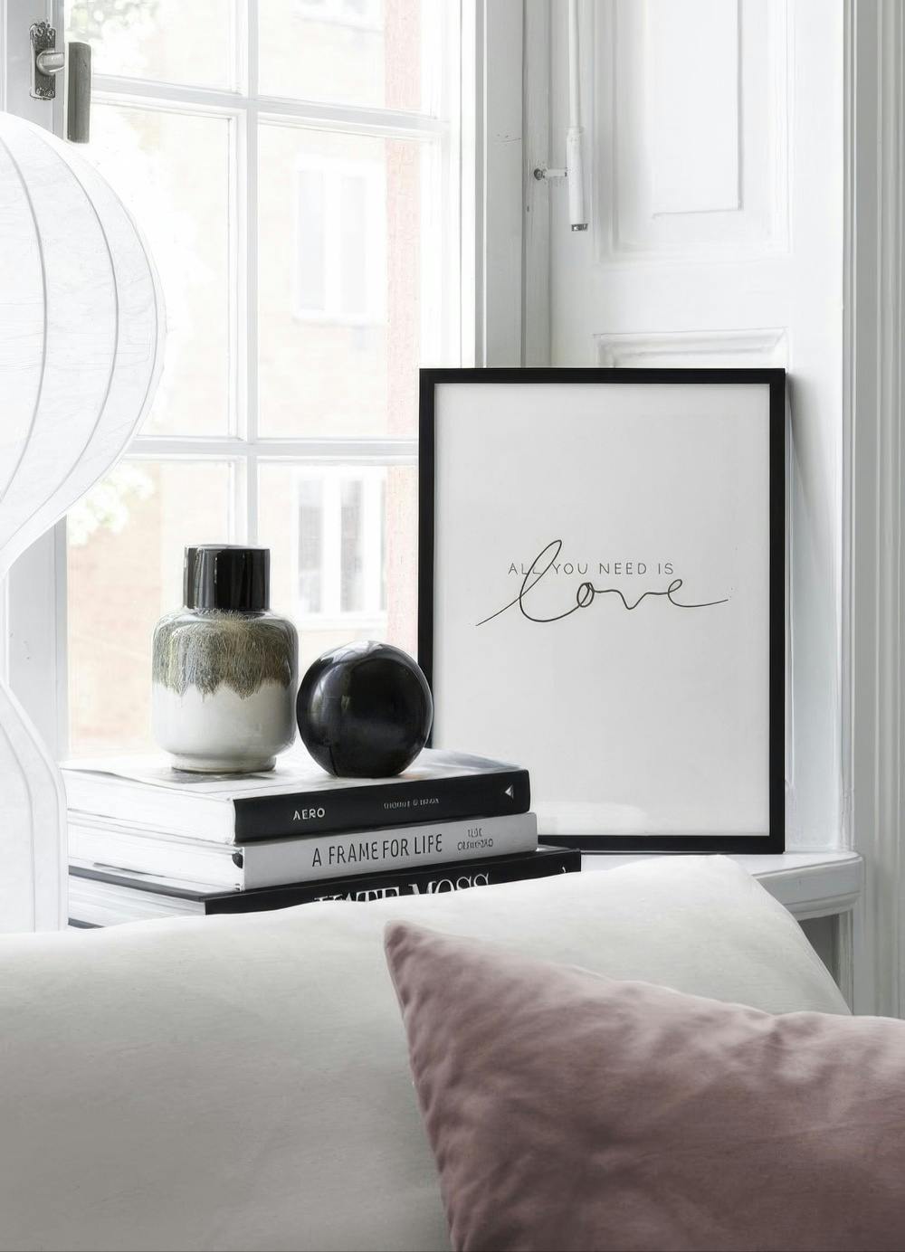 Affiche All you need is love avec une typographie manuscrite, posée sur une pile de livres près dune fenêtre.