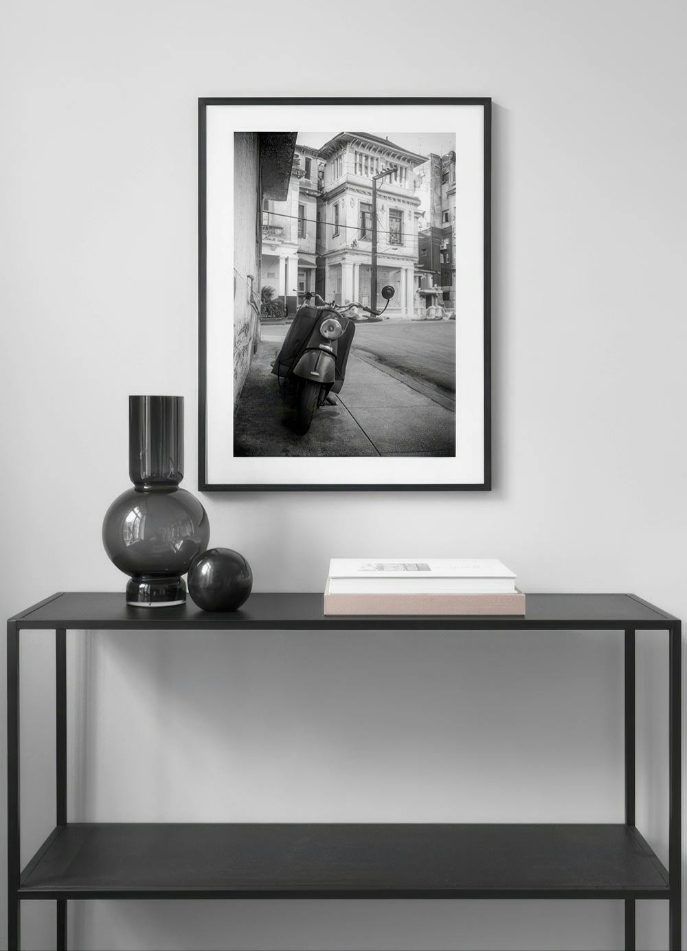 Affiche noir et blanc dun scooter garé dans la rue, devant un bâtiment ancien, affichée au-dessus dune console.