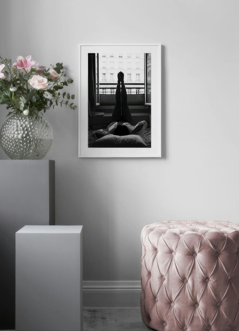 Affiche noir et blanc dune personne allongée, jambes levées, face à une fenêtre urbaine, dans un salon moderne.