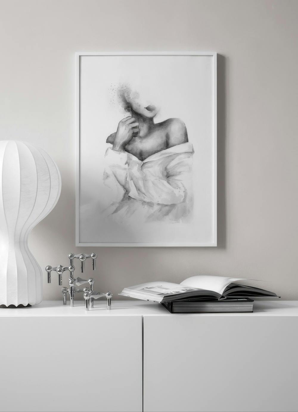 Affiche dune femme en chemise blanche, le visage estompé, accrochée au-dessus dun meuble blanc avec lampe et livres.
