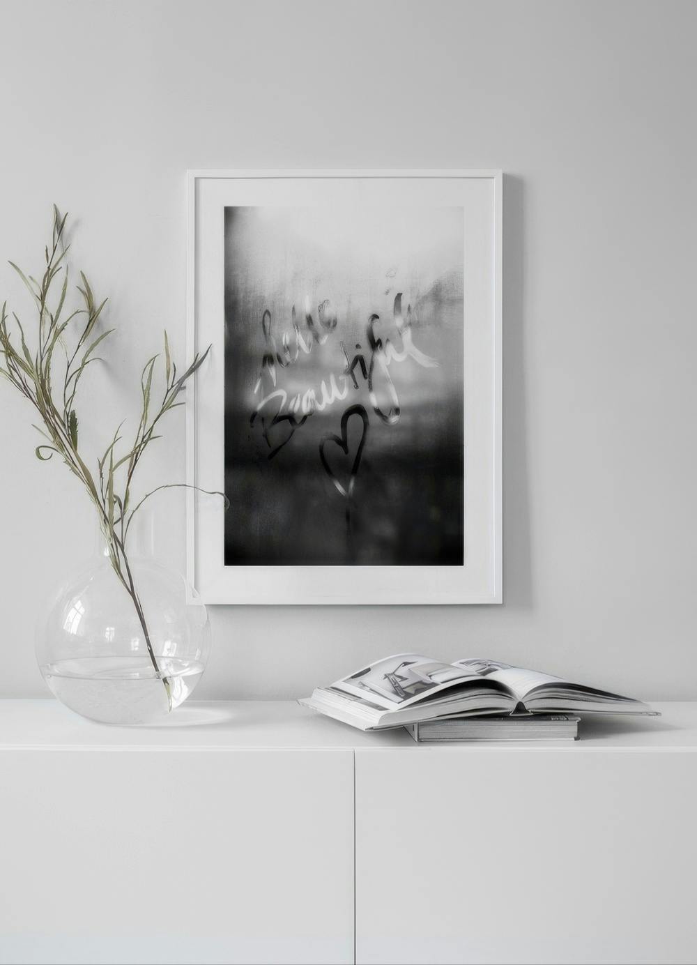 Affiche Hello Beautiful avec un cœur sur fond noir et blanc, accrochée au-dessus dun meuble blanc avec un vase et un livre.