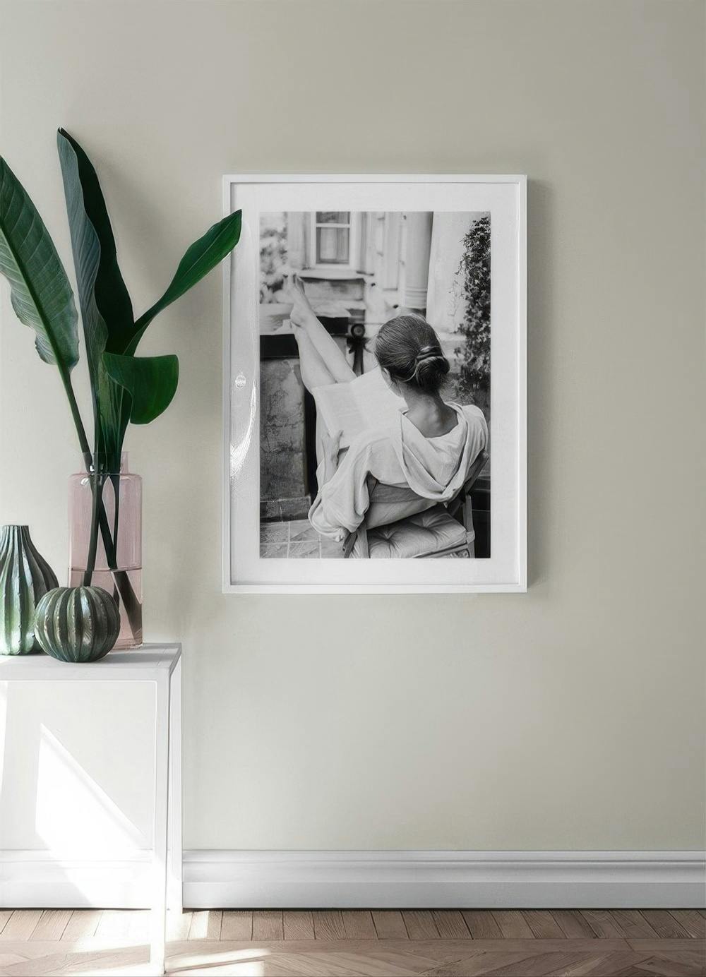 Affiche en noir et blanc dune femme lisant, jambes relevées, dans un intérieur lumineux avec plante et vases.