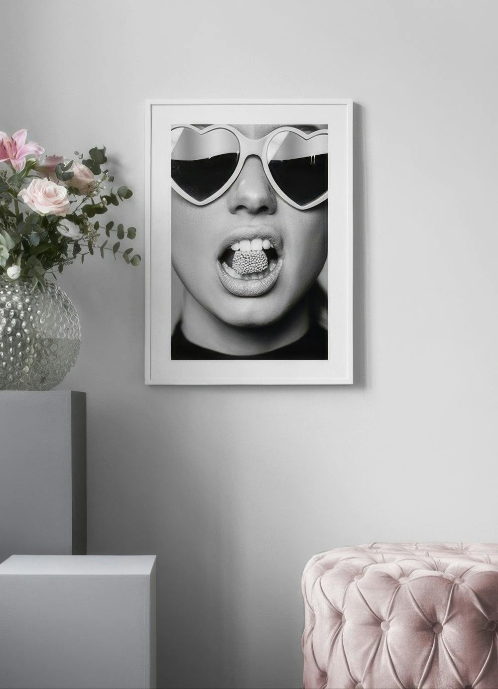 Affiche noir et blanc dun visage féminin avec des lunettes cœur et des bonbons dans la bouche, sur un mur gris avec un vase de f