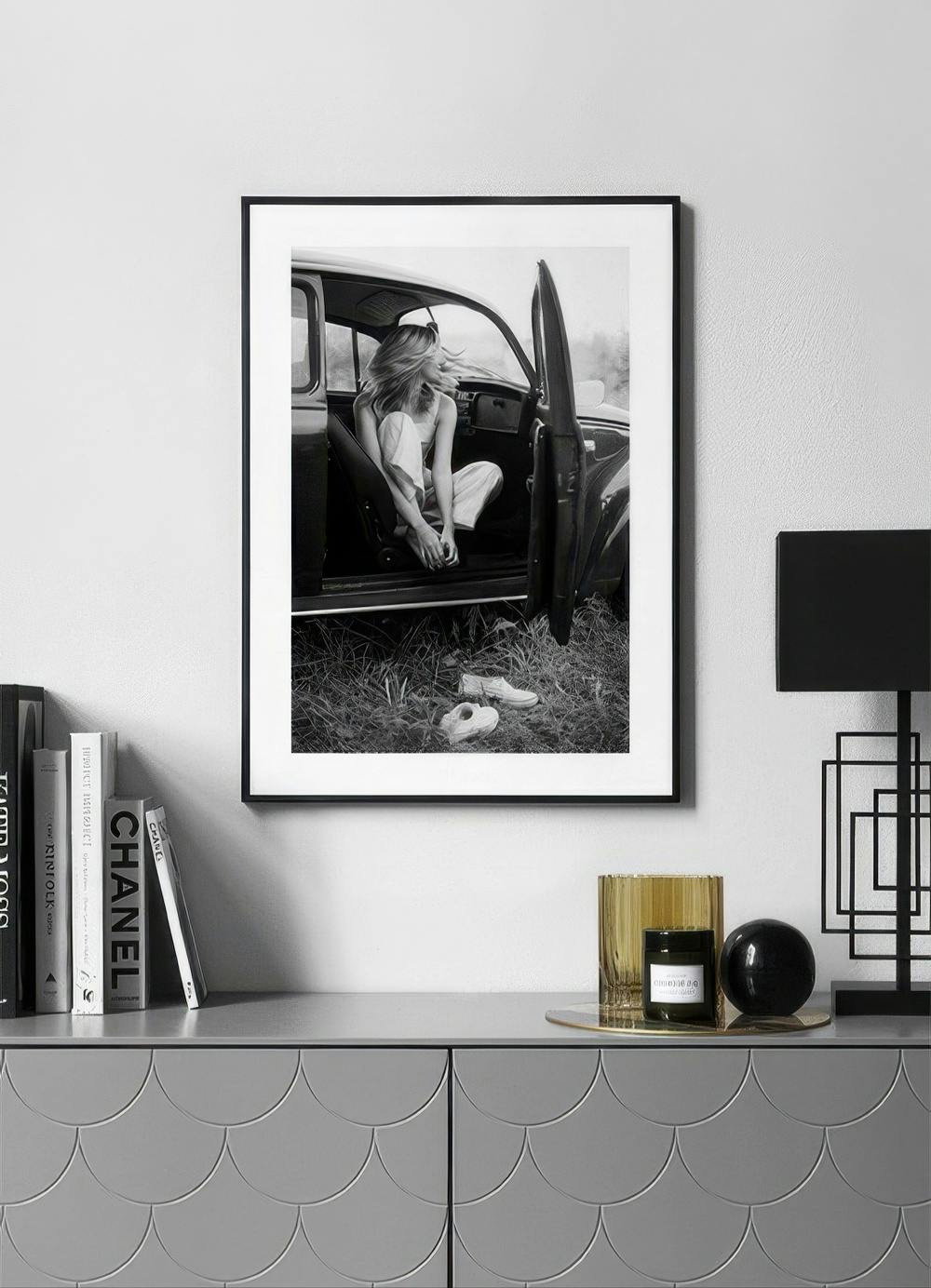 Affiche noir et blanc dune femme assise dans une voiture, exposée au-dessus dune commode grise moderne.