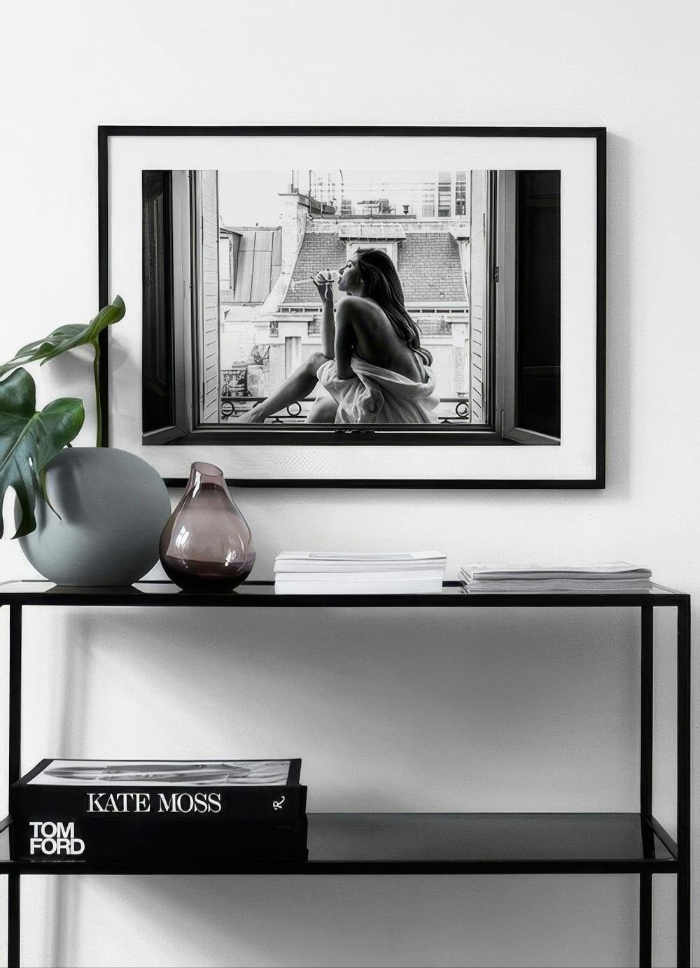 Affiche noir et blanc dune femme buvant, assise à une fenêtre avec vue sur les toits de Paris, au-dessus dune console noire.