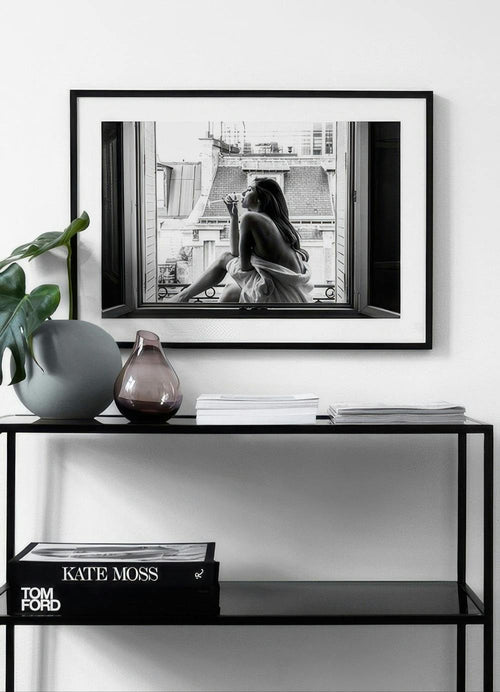 Affiche noir et blanc dune femme buvant, assise à une fenêtre avec vue sur les toits de Paris, au-dessus dune console noire.