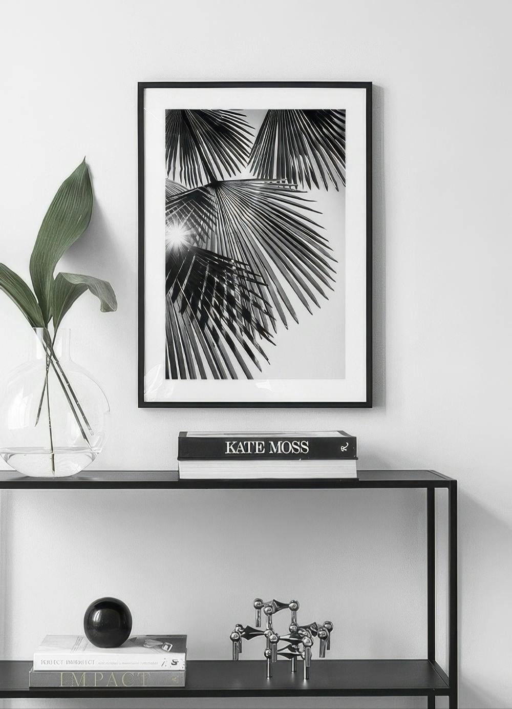Affiche noir et blanc de palmiers sous le soleil, accrochée au-dessus dune étagère noire avec des livres et une plante.