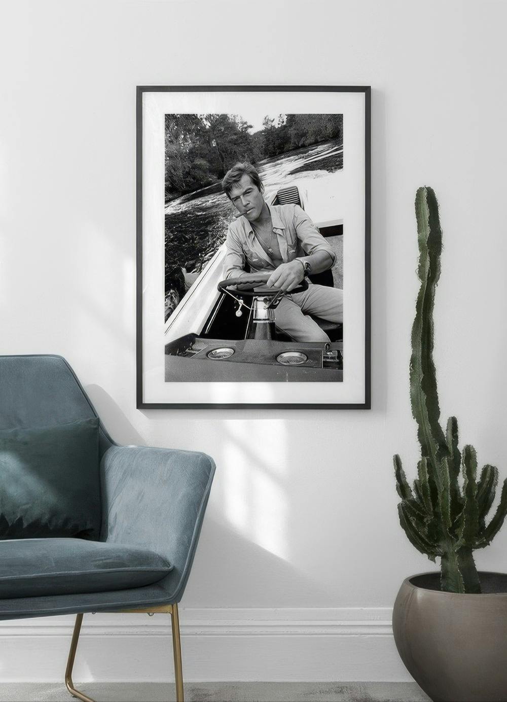 Affiche en noir et blanc dun homme en bateau, accrochée au-dessus dun fauteuil bleu et dun grand cactus.