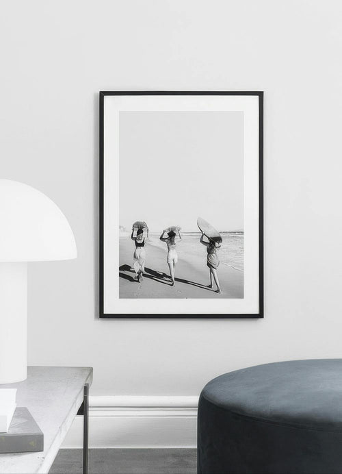 Affiche en noir et blanc de trois femmes avec des planches de surf sur la plage, affichée dans un salon moderne.