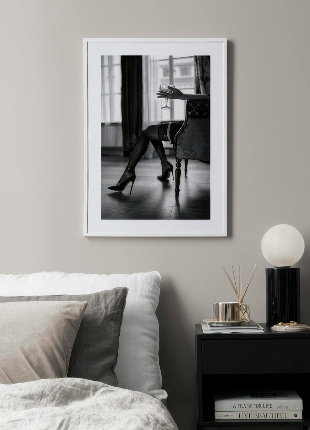Affiche noir et blanc dune femme en talons et bas, assise sur un fauteuil, tenant un verre. Accrochée dans une chambre moderne a