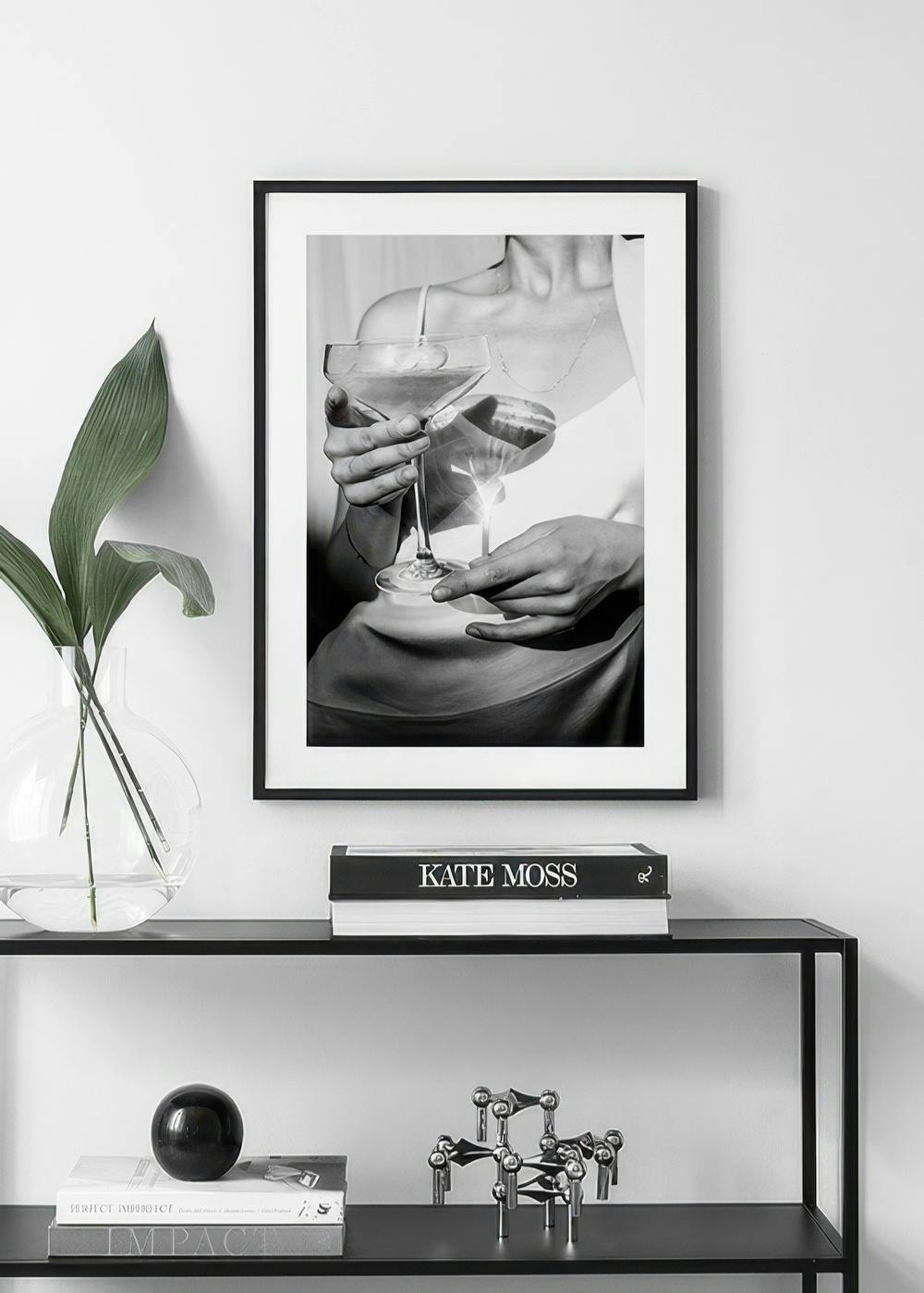 Affiche en noir et blanc dune femme tenant un verre à cocktail, accrochée au-dessus dune étagère noire avec des livres et une pl