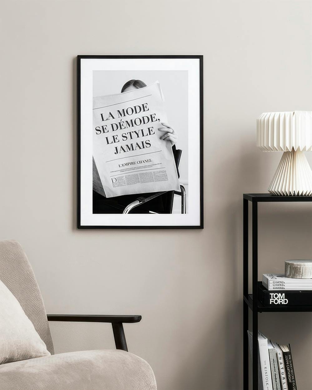 Affiche noir et blanc dune personne lisant un journal La Mode Se Démodé..., sur un mur clair à côté dun fauteuil et dune étagère