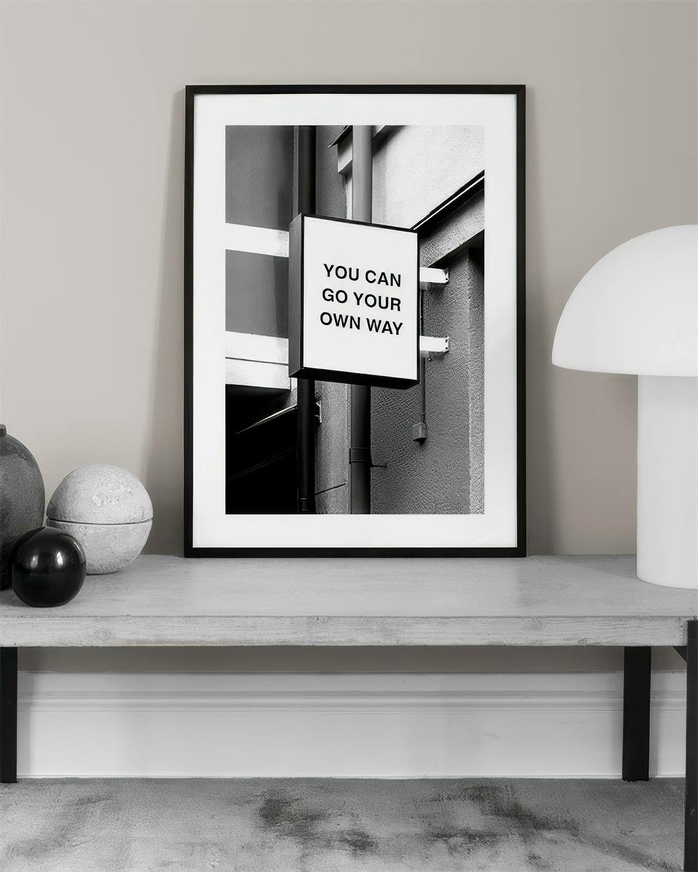 Affiche noir et blanc avec un panneau YOU CAN GO YOUR OWN WAY dans un espace urbain, posée sur un banc.