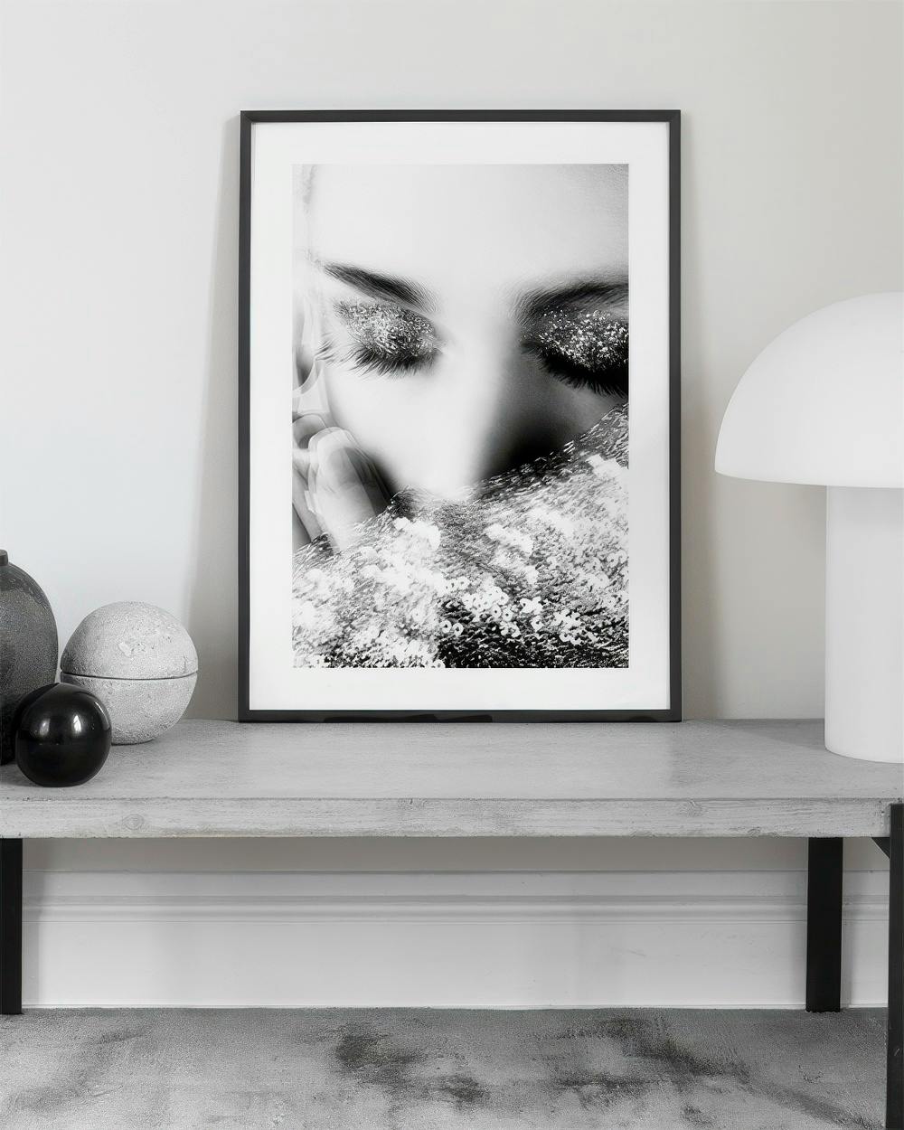 Affiche noir et blanc dun visage féminin aux paupières pailletées, posée sur une console en béton.