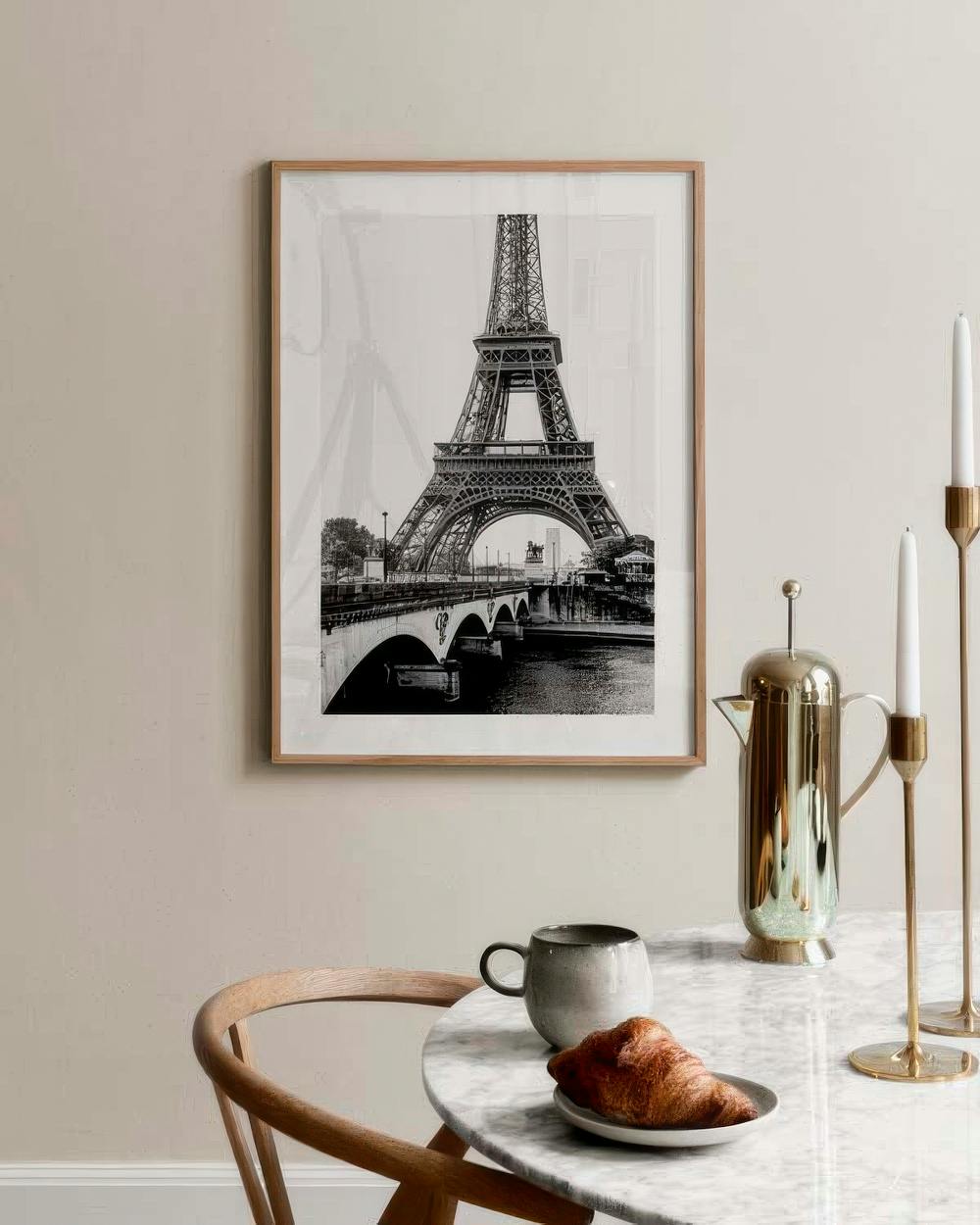 Affiche noir et blanc de la Tour Eiffel et dun pont sur la Seine, accrochée au-dessus dune table avec tasse et croissant.
