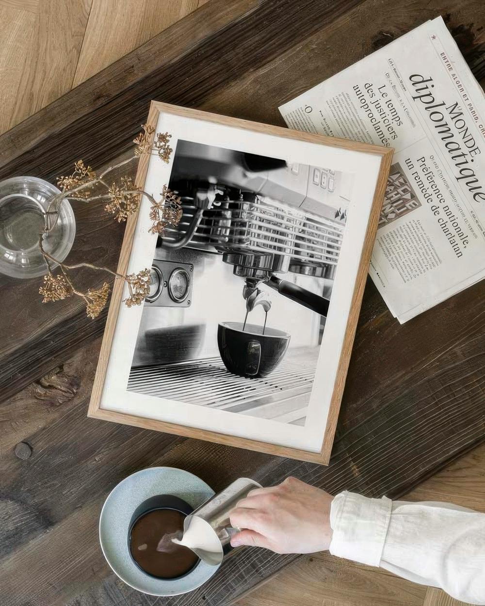 Affiche en noir et blanc dune machine à café versant un expresso, posée sur une table en bois avec un journal.