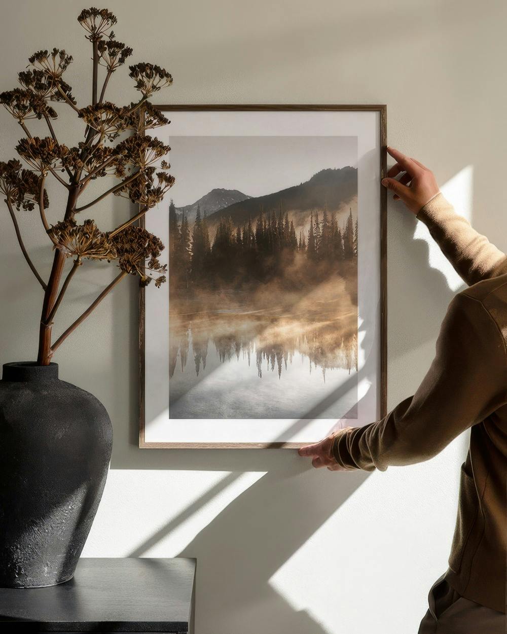 Affiche de forêt brumeuse et montagnes enneigées au-dessus dun meuble noir avec un vase, installée par une personne.
