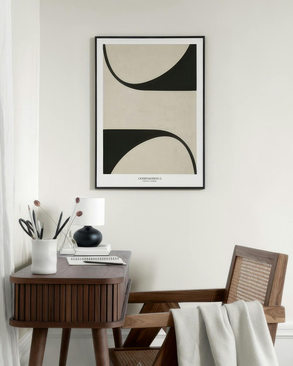 Affiche avec des formes organiques noires et beiges, accrochée au mur au-dessus dune table de chevet et dun fauteuil en bois.