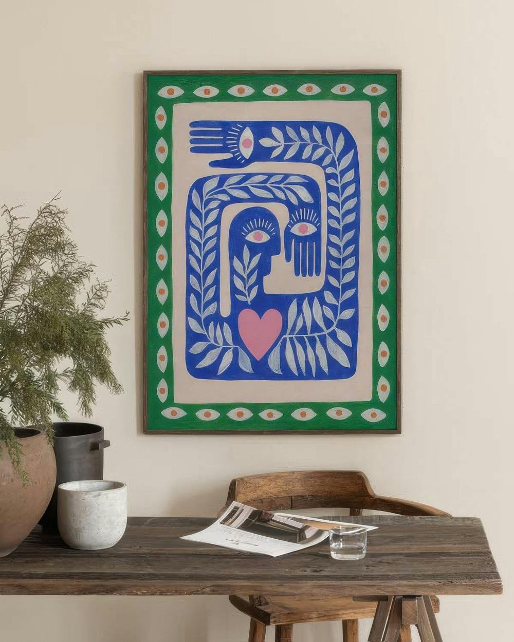 Affiche avec un visage et des mains bleues, un cœur rose et des motifs de feuilles, accrochée au-dessus dun bureau en bois.
