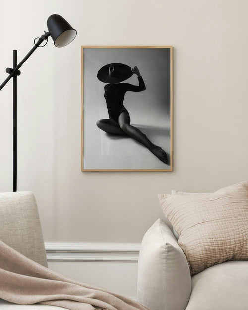 Affiche noir et blanc dune femme assise avec un chapeau, suspendue au-dessus dun canapé beige dans un salon.