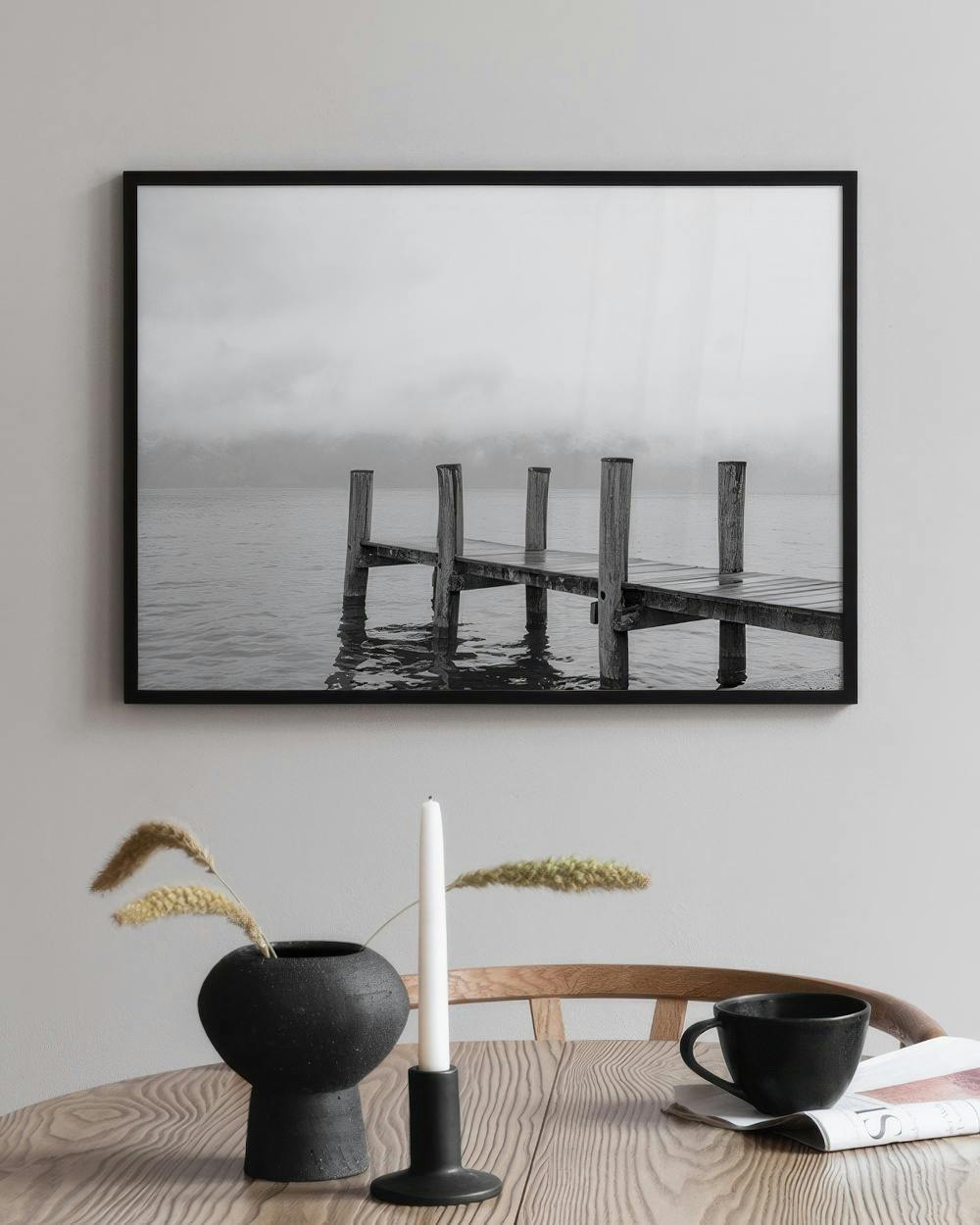 Affiche en noir et blanc d’un ponton sur un lac brumeux, accrochée au-dessus d’une table en bois avec vase et bougie.