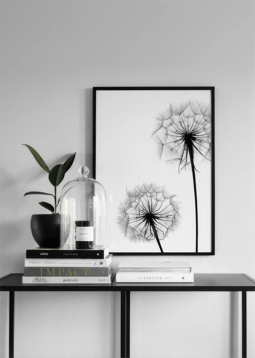 Affiche en noir et blanc de deux pissenlits sur un mur gris, avec une plante et des livres sur une table.