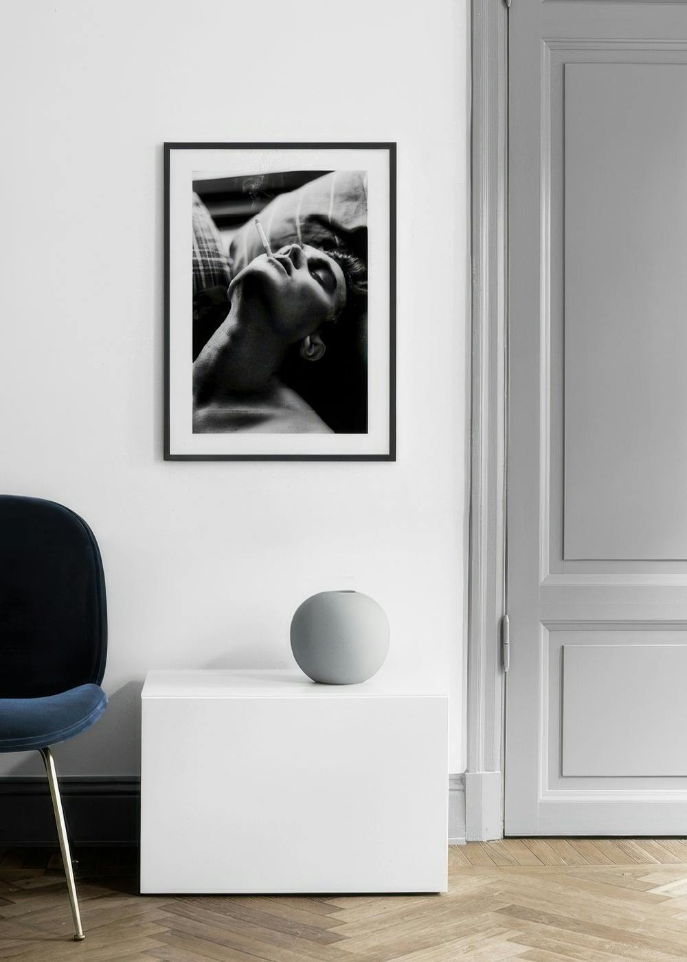Affiche noir et blanc dun homme fumant, suspendue au-dessus dun meuble blanc avec un vase rond gris, à côté dune porte.