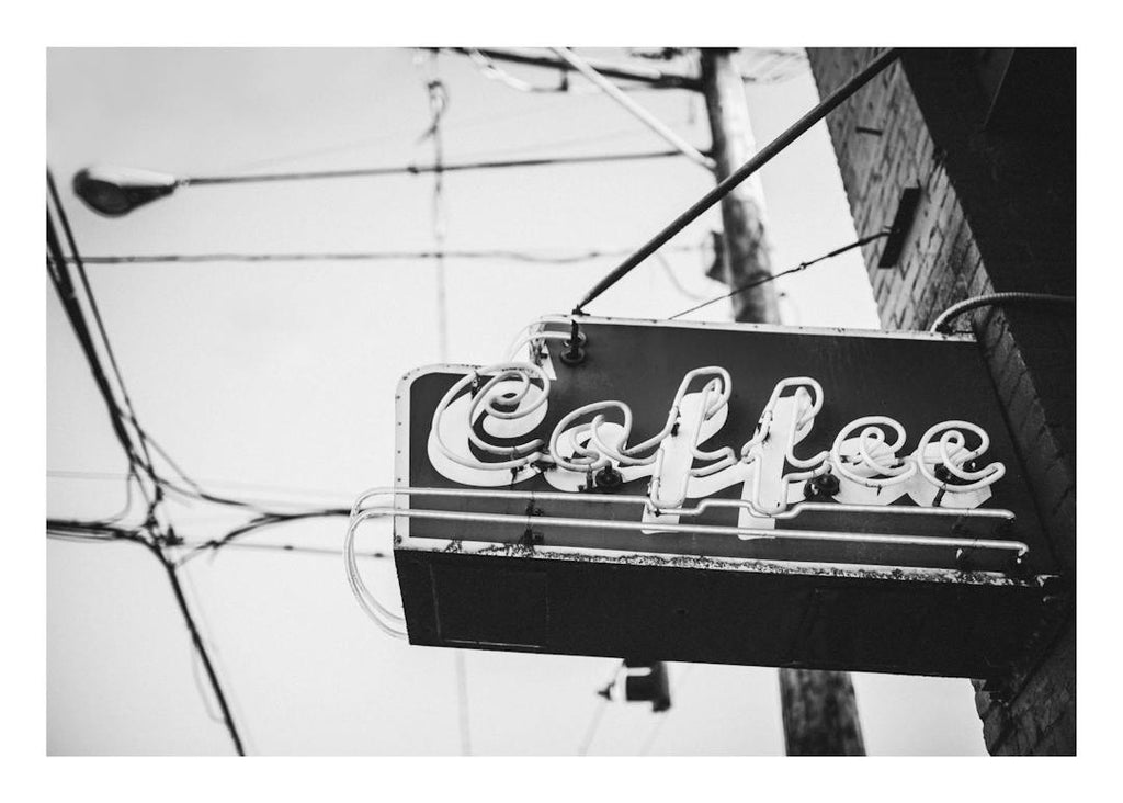 Affiche noir et blanc dune enseigne Coffee au typographie néon sur un bâtiment en briques.
