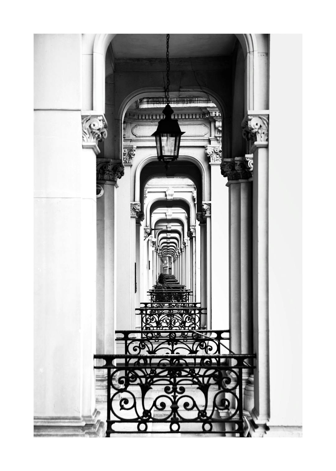 Affiche noir et blanc dune longue galerie darches et de balustrades en fer forgé, créant une perspective infinie.