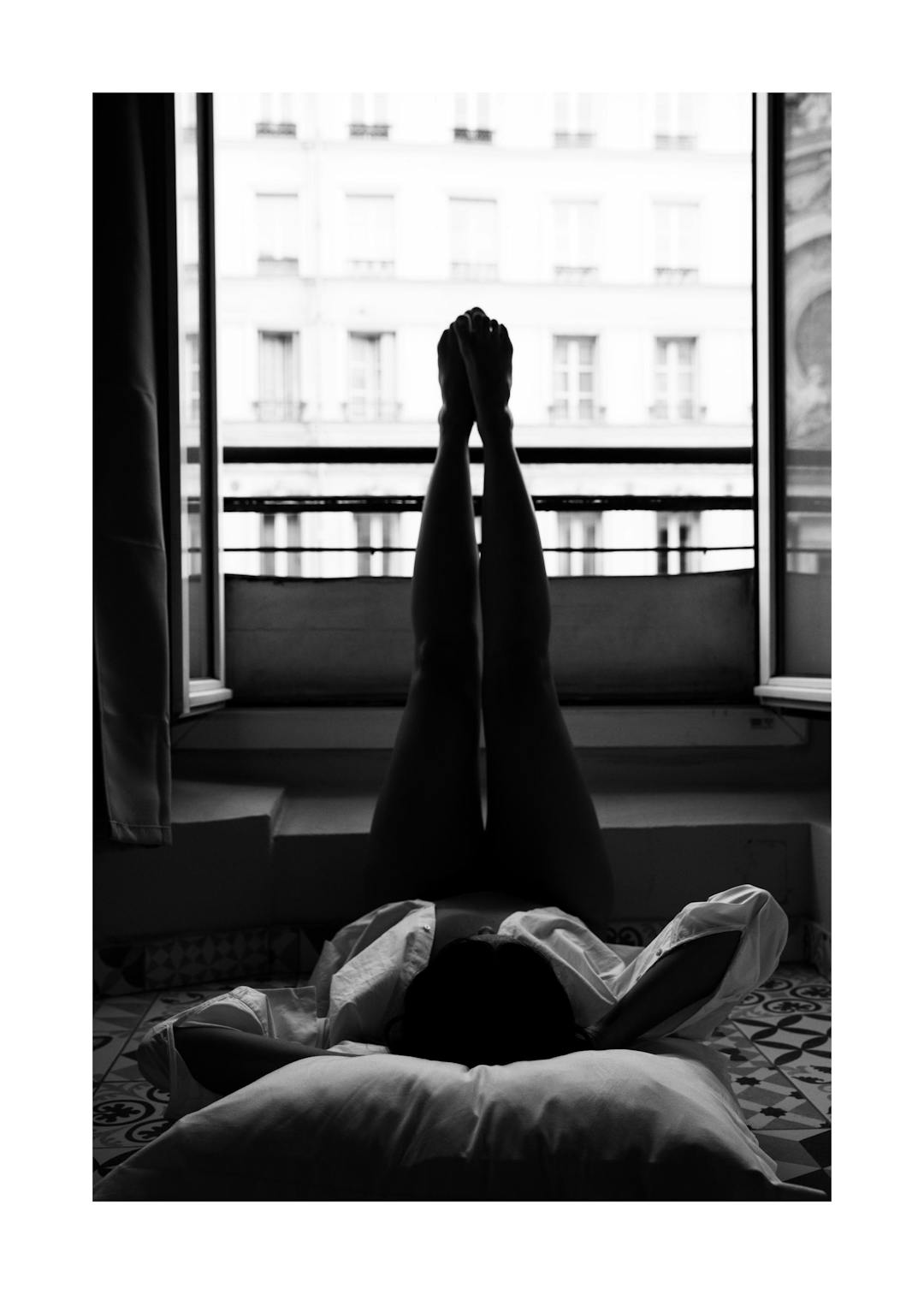 Affiche noir et blanc dune personne allongée, jambes levées vers une fenêtre parisienne avec des immeubles en arrière-plan.