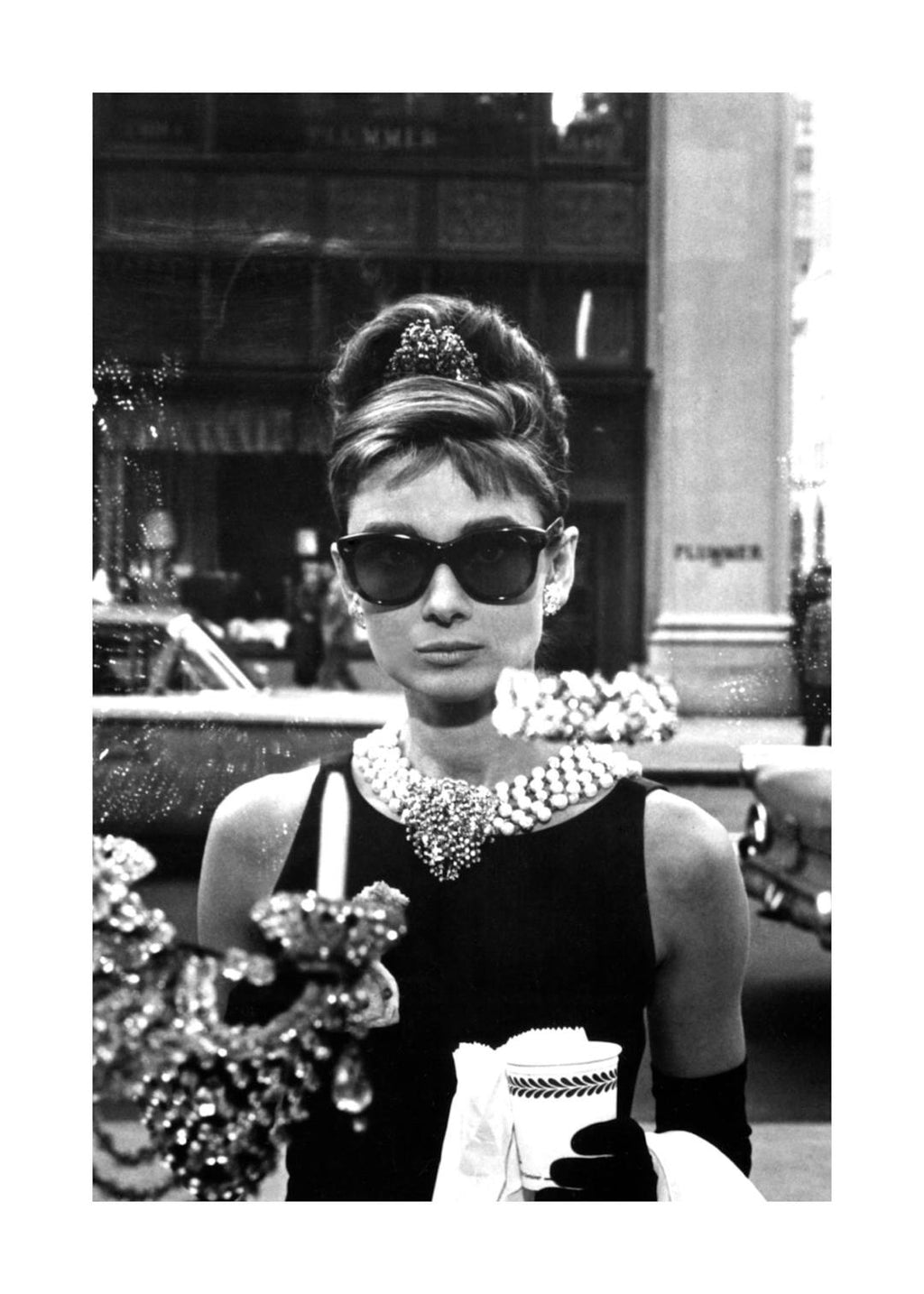 Affiche noir et blanc dAudrey Hepburn devant une vitrine, portant des lunettes de soleil et tenant une tasse de café.