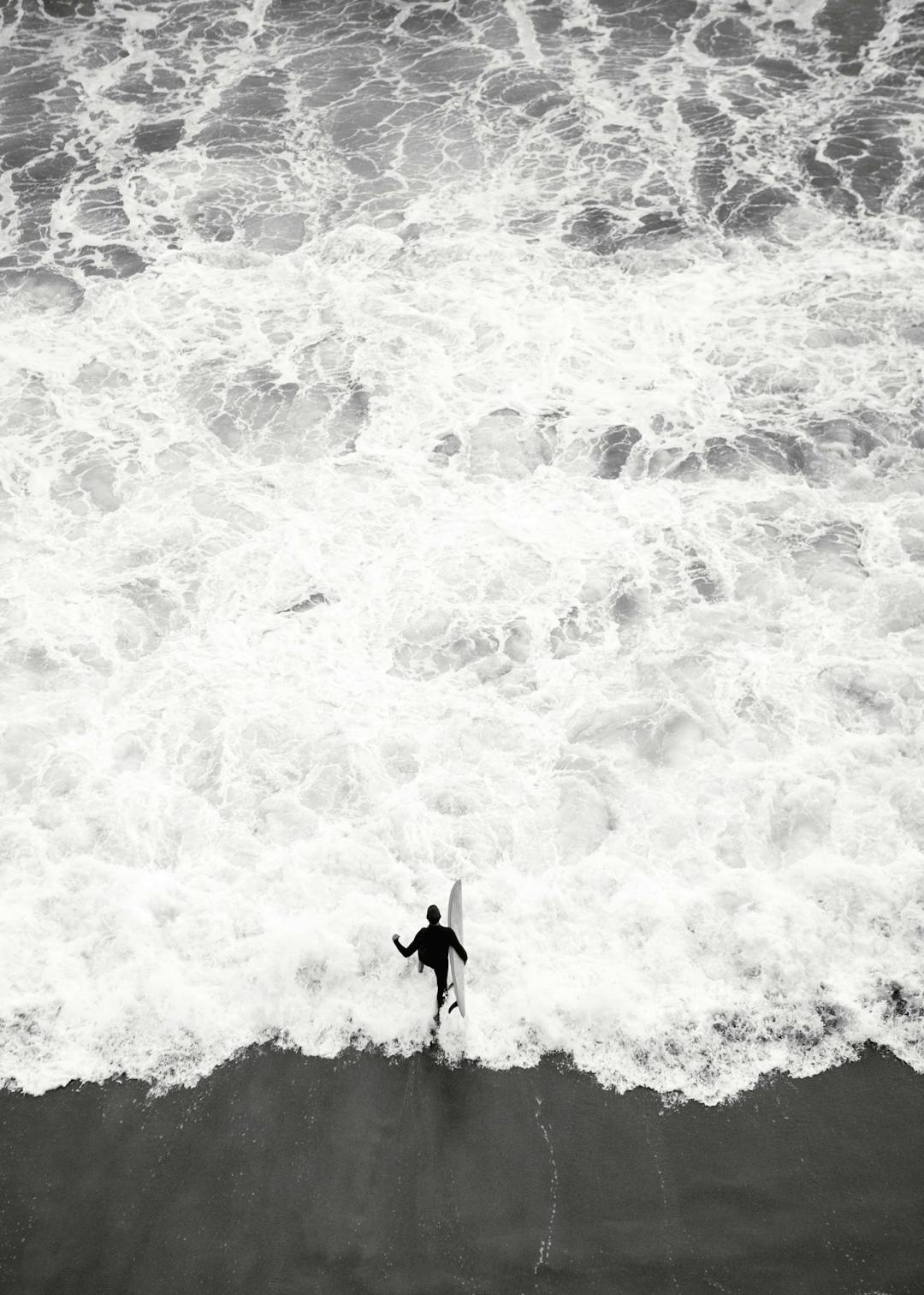 Affiche noir et blanc. Un surfeur avec une planche entre dans locéan, des vagues écumantes en arrière-plan.