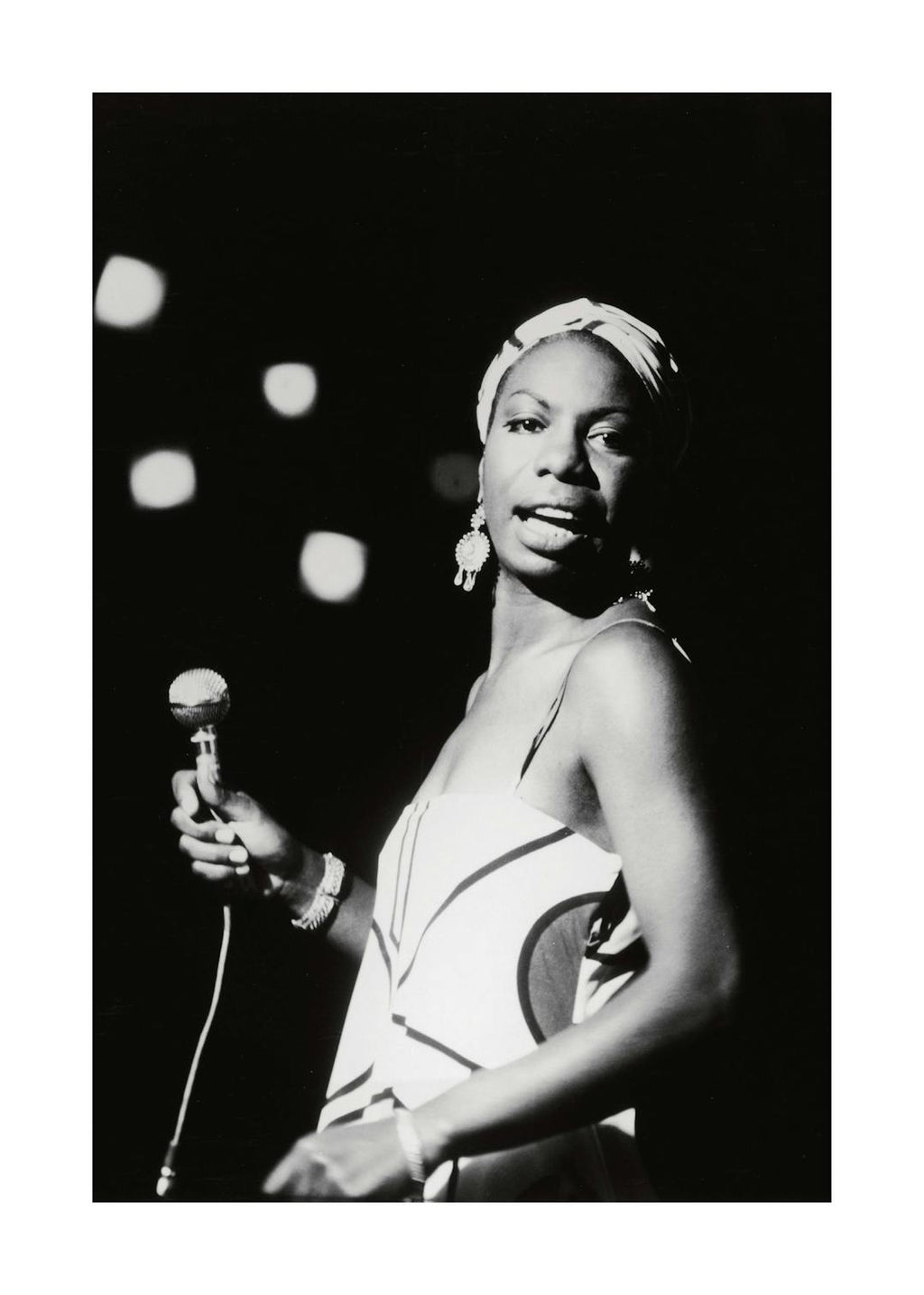 Affiche noir et blanc de Nina Simone tenant un micro, avec un foulard et des boucles doreilles, sur scène.