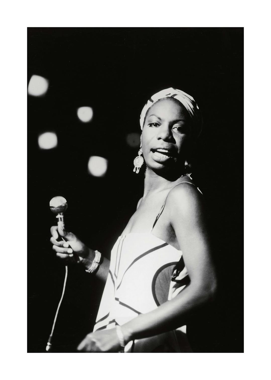 Affiche noir et blanc de Nina Simone tenant un micro, avec un foulard et des boucles doreilles, sur scène.