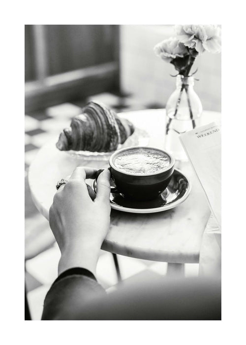 Affiche noir et blanc dune main tenant une tasse de café sur une table de café, avec un croissant et un journal