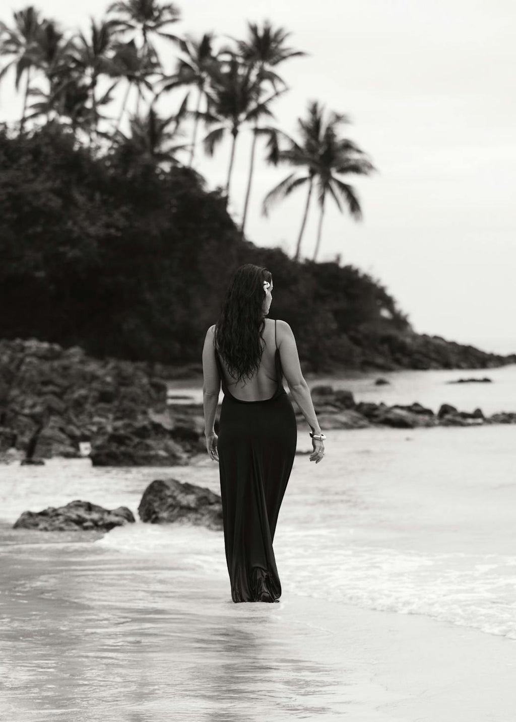Affiche en noir et blanc dune femme en robe longue marchant sur une plage tropicale avec des palmiers.