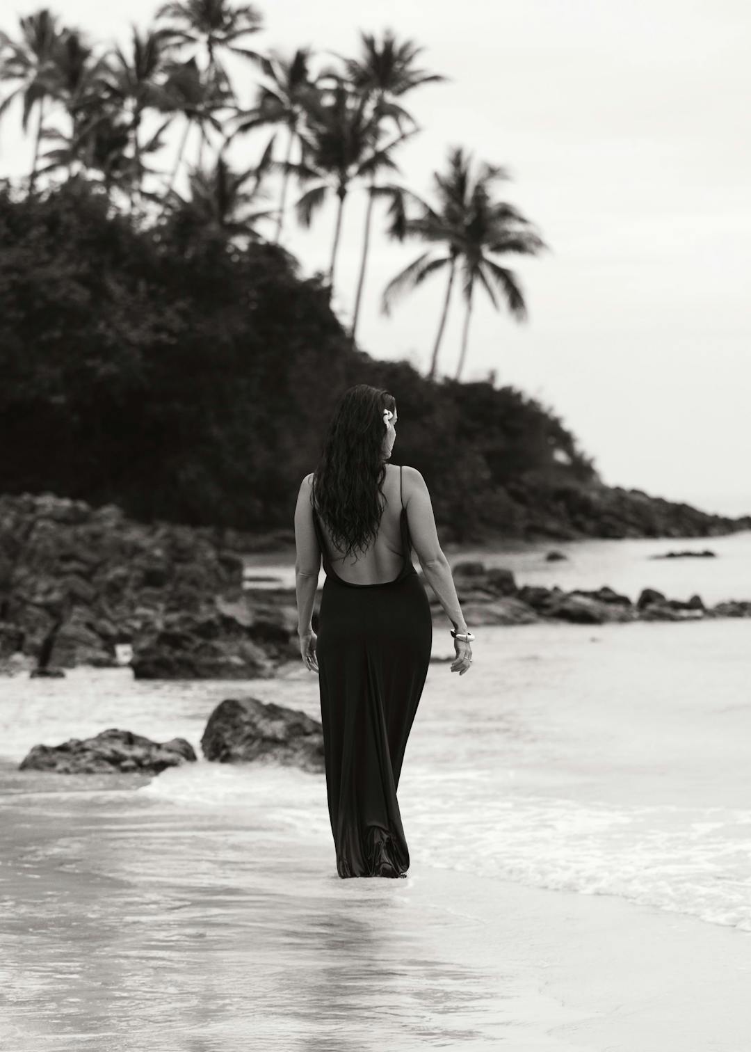 Affiche en noir et blanc dune femme en robe longue marchant sur une plage tropicale avec des palmiers.
