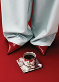 Affiche dune personne en pantalon gris clair et chaussures à talons rouges, avec une tasse de café argentée au premier plan sur 