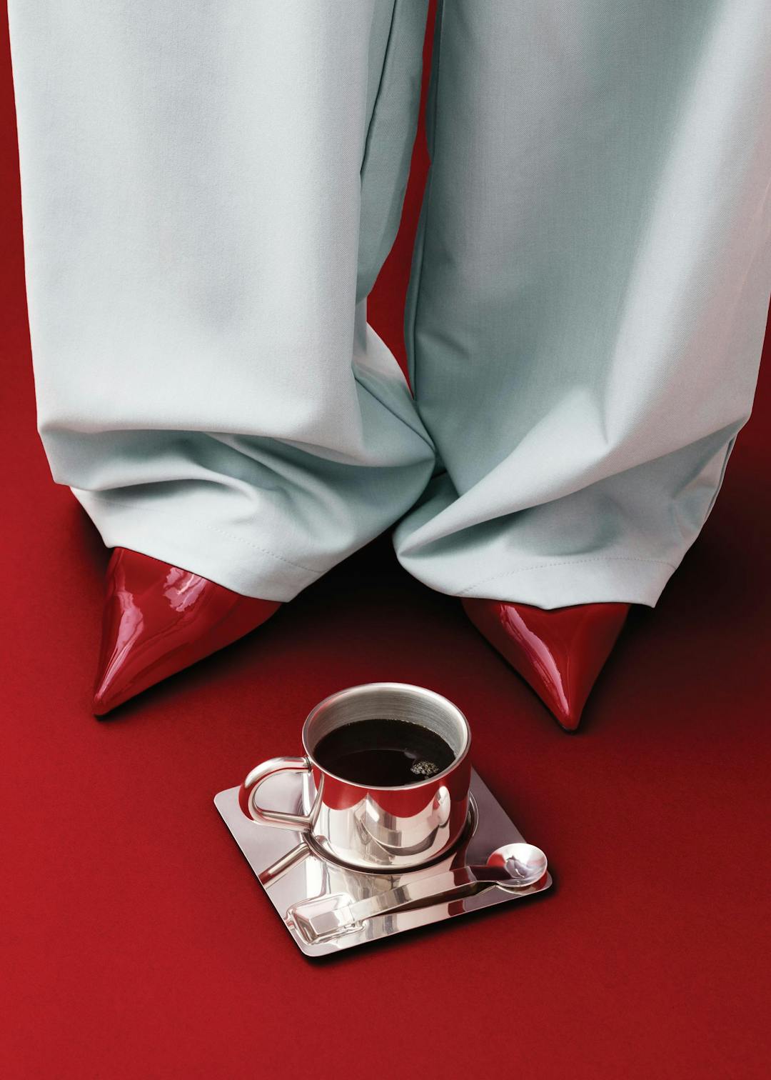 Affiche dune personne en pantalon gris clair et chaussures à talons rouges, avec une tasse de café argentée au premier plan sur 