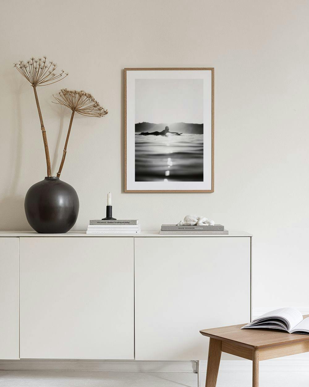 Affiche noir et blanc dun surfeur couché sur une planche au lever du soleil, accrochée au mur près dun fauteuil et dun cactus.
