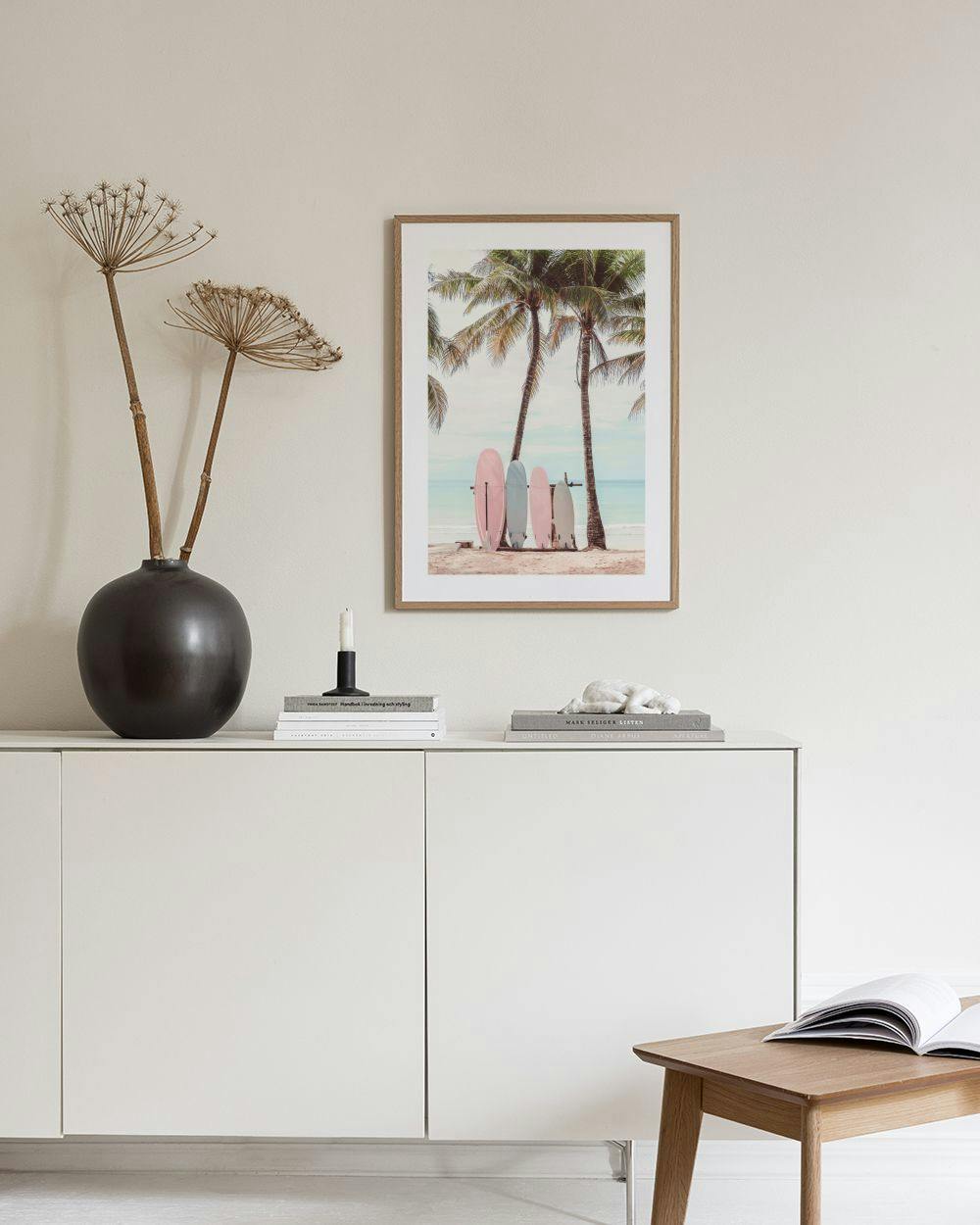 Affiche de planches de surf pastel sur une plage, avec palmiers, posée sur un meuble blanc avec vase et livre.