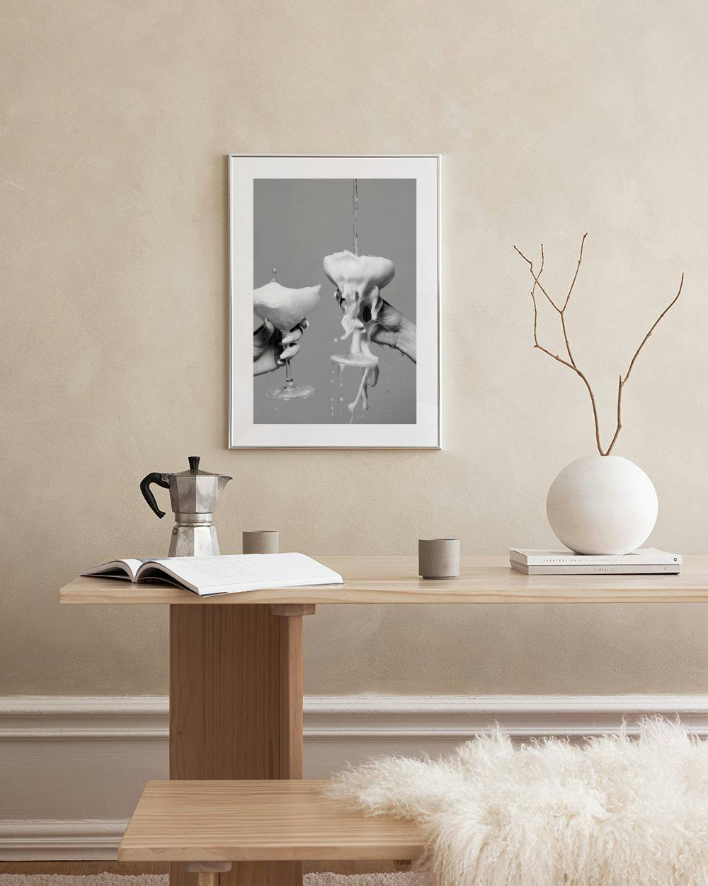 Affiche noire et blanche de mains tenant des coupes débordant de mousse, suspendue au-dessus dun meuble blanc avec un vase gris.