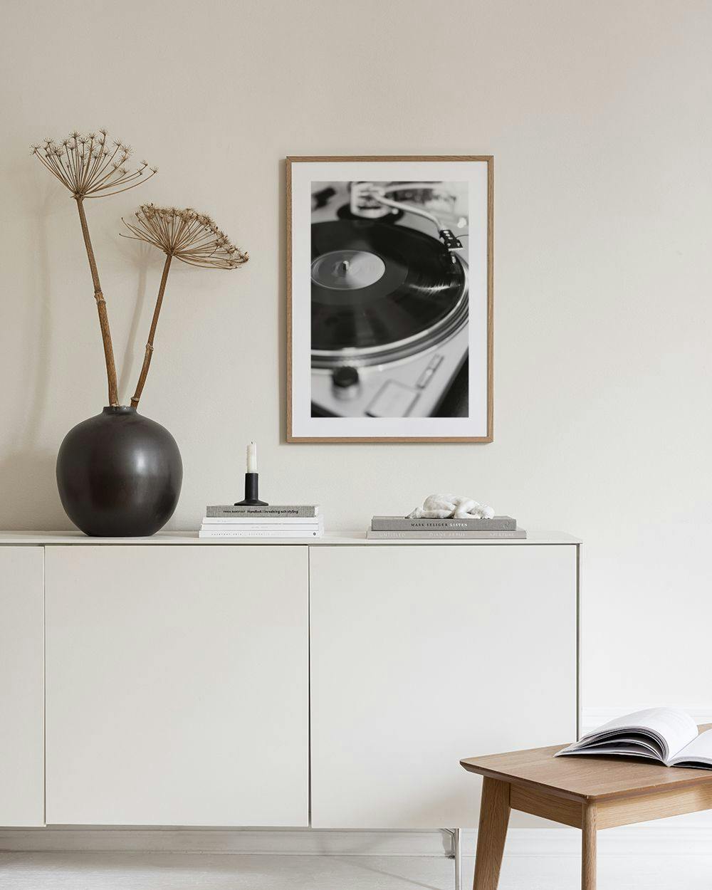 Affiche noir et blanc dune platine vinyle avec un disque, posée sur une console grise avec des objets décoratifs et une lampe.